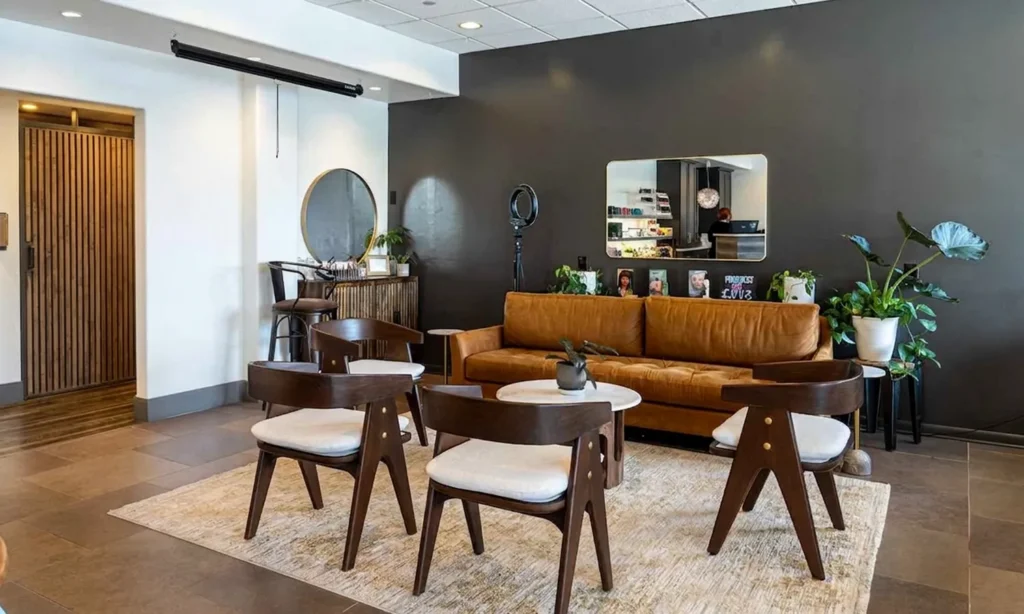 - Bella Capelli Sanctuario in Westlake, OH Modern lounge area with a brown leather sofa, four wooden chairs, a beige rug, a round mirror, and various potted plants against a dark accent wall. - Bella Capelli Sanctuario in Westlake, OH