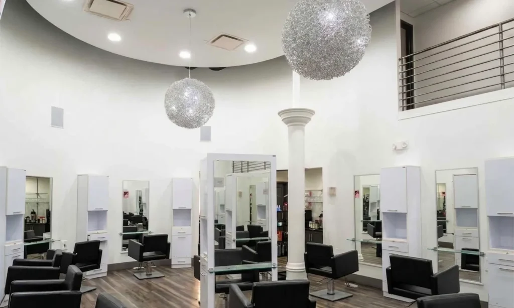 - Bella Capelli Sanctuario in Westlake, OH A modern hair salon interior with black chairs, white stations, large spherical pendant lights, and a high ceiling. There is a staircase and railing visible on the upper right. - Bella Capelli Sanctuario in Westlake, OH