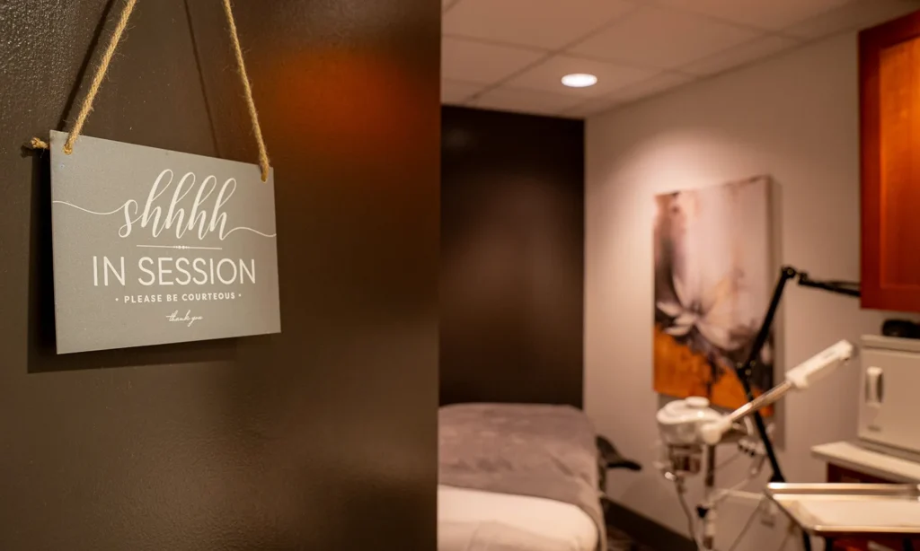 - Bella Capelli Sanctuario in Westlake, OH A "Shhh, In Session. Please Be Courteous. Thank You" sign hangs on a dark wall in a room with a massage table, spa equipment, and artwork. - Bella Capelli Sanctuario in Westlake, OH