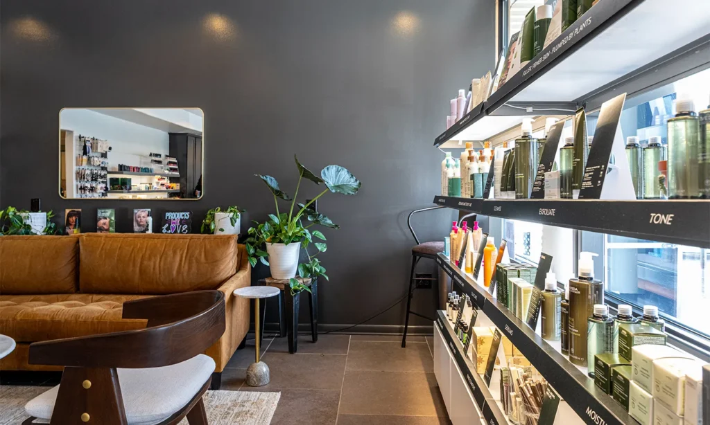 - Bella Capelli Sanctuario in Westlake, OH A modern salon interior with a brown sofa, indoor plants, shelves stocked with hair products, and a large wall-mounted mirror. - Bella Capelli Sanctuario in Westlake, OH