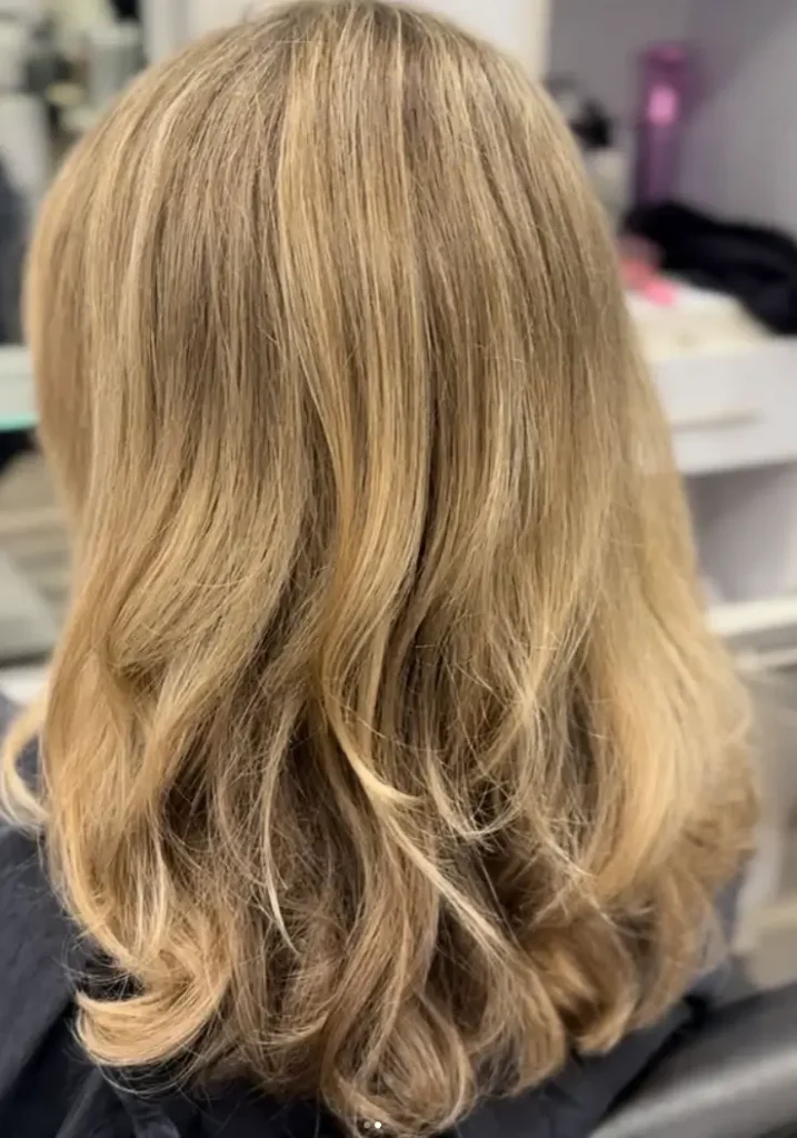- Bella Capelli Sanctuario in Westlake, OH A person with shoulder-length, wavy blonde hair sits in a salon chair, facing away from the camera. - Bella Capelli Sanctuario in Westlake, OH
