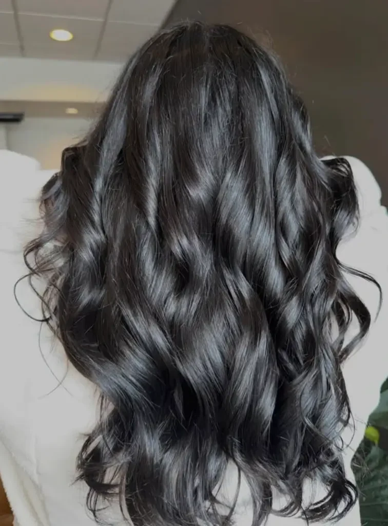- Bella Capelli Sanctuario in Westlake, OH A person with long, dark, wavy hair is shown from the back, wearing a light-colored top indoors. - Bella Capelli Sanctuario in Westlake, OH
