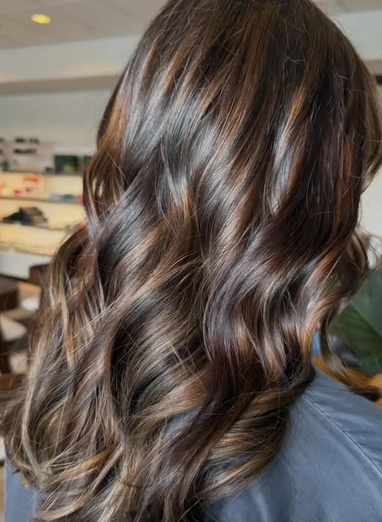 - Bella Capelli Sanctuario in Westlake, OH Person with long, wavy, dark brown hair featuring caramel highlights, seen from the back in an indoor setting. - Bella Capelli Sanctuario in Westlake, OH